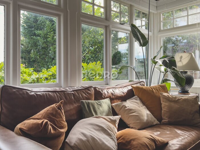 Papier peint  A brown leather sofa with scattered pillows, typical windows and a strelitzia plant in atypical British home. Interior decor.