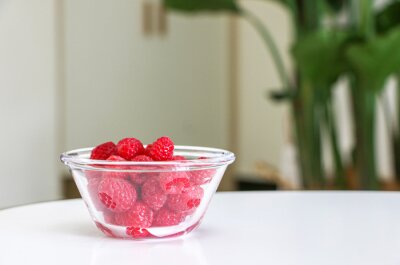 Papier peint  A glass bowl of juicy raspberries on white table of living room interior, cupboards, drawers, and Giant White Bird of Paradise plant (Strelitzia nicolai) out of focus in background, healthy lifestyle