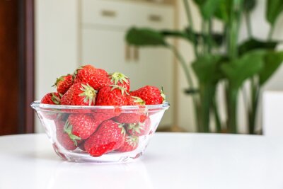 Papier peint  A glass bowl of juicy red strawberries placed on white table of living room interior, cupboards, drawers, and Giant White Bird of Paradise plant (Strelitzia nicolai) out of focus in background