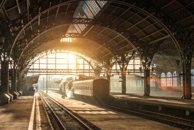 Papier peint  Ancienne gare avec un train et une locomotive sur le quai en attente de départ. Les rayons du soleil du soir dans les arcs de fumée.