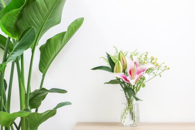 Papier peint  Beautiful plants and flowers decorating home interior, bouquet of flowers (pink lilies and daisies) in glass vase on wooden surface next to Giant White Bird of Paradise Plant (Strelitzia nicolai)