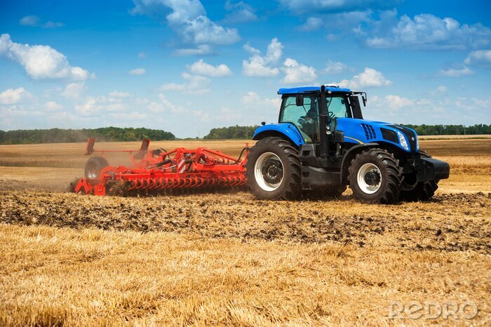 Papier peint  blue new tractor pulls a red harrow raises dust at the field, soil preparation