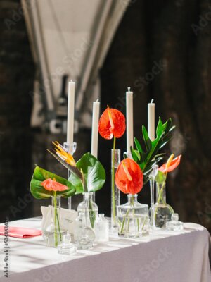 Papier peint  Bouquets of red anthurium, strelitzia, and tropical leaves stand in glass vases on a white table. There are white candles in the background. Tropical decor.