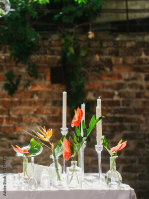 Papier peint  Bouquets of red anthurium, strelitzia, and tropical leaves stand in glass vases on a white table. There are white candles in the background. Tropical decor.