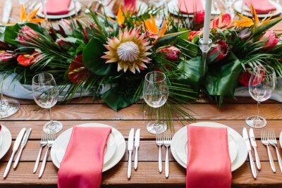 Papier peint  Decor and serving of a wedding banquet in a tropical style. Exotic flowers, protea, strelitzia, leaves, white candles in high candlesticks, wine glasses, red napkins on white plates, cutlery on a wood