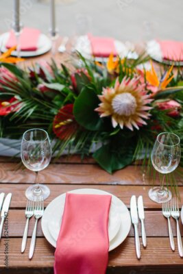 Papier peint  Decor and serving of a wedding banquet in a tropical style. Exotic flowers, protea, strelitzia, leaves, white candles in high candlesticks, wine glasses, red napkins on white plates, cutlery on a wood