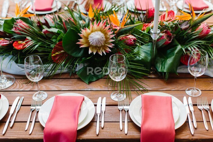 Papier peint  Decor and serving of a wedding banquet in a tropical style. Exotic flowers, protea, strelitzia, leaves, white candles in high candlesticks, wine glasses, red napkins on white plates, cutlery on a wood