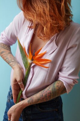 Papier peint  fille dans une chemise rose posant avec une fleur strelitzia dans son tatouage les mains sur un fond bleu