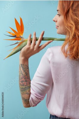Papier peint  Girl with a tattoo holding on her shoulder an orange flower strelitzia around a blue background with space for text. Creative layout