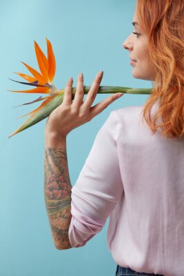 Papier peint  Girl with a tattoo holding on her shoulder an orange flower strelitzia around a blue background with space for text. Creative layout