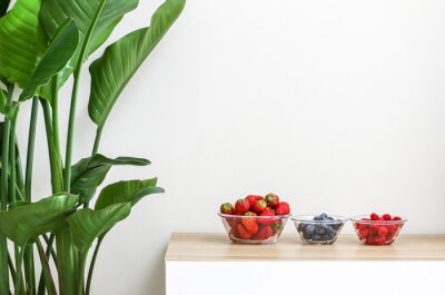 Papier peint  Glass bowls of juicy and fresh strawberries, blueberries and raspberries on wooden table next to a beautiful Giant White Bird of Paradise Plant (Strelitzia nicolai), freshening and decorating home