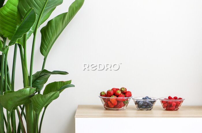 Papier peint  Glass bowls of juicy and fresh strawberries, blueberries and raspberries on wooden table next to a beautiful Giant White Bird of Paradise Plant (Strelitzia nicolai), freshening and decorating home