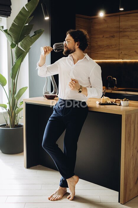 Papier peint  Handsome curly man at home in a beautiful kitchen drinks red wine and tastes cheese and olives.On the background of a beautiful strelitzia flower