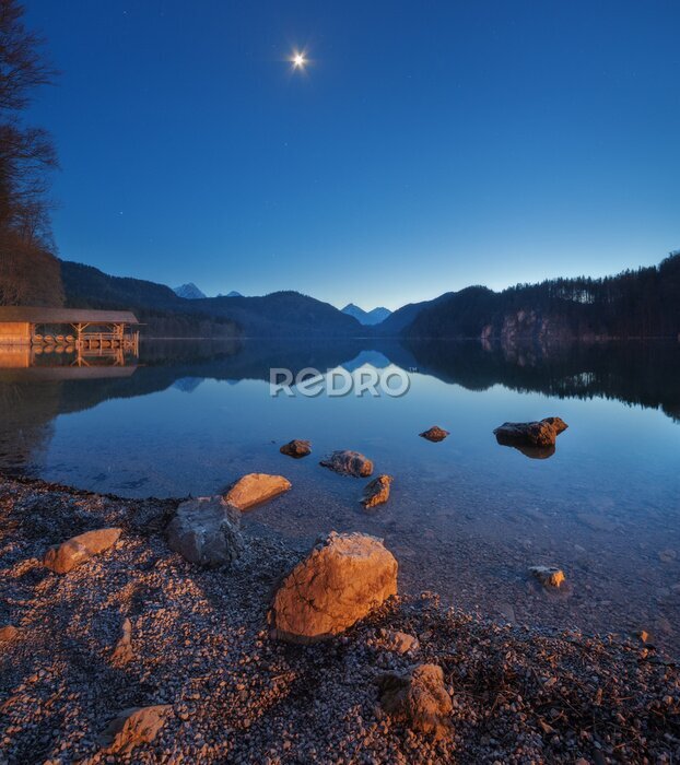 Papier peint  Nuit dans le lac d'Alpsee en Allemagne. Beau paysage avec le lac, les montagnes, la forêt, les étoiles, la pleine lune, le ciel bleu et les pierres dans l'eau. Photo panoramique. Printemps