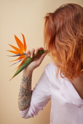 Papier peint  Strelitzia flower decorating the shoulder of a red-haired girl with a tattoo on a beige background with space for text. Creative layout
