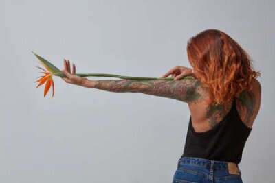 Papier peint  Stylish red-haired girl with a tattoo from back holds a strelitzia flower around a gray background. Photo as a layout for your ideas