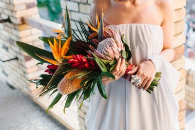 Papier peint  The bride in a dress with bare shoulders holds an exotic wedding bouquet of tropical flowers. Bouquet of protea, strelitzia, red ginger, and tropical leaves. In the background is a brick wall.