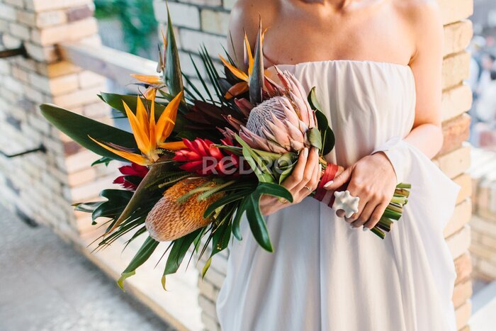 Papier peint  The bride in a dress with bare shoulders holds an exotic wedding bouquet of tropical flowers. Bouquet of protea, strelitzia, red ginger, and tropical leaves. In the background is a brick wall.