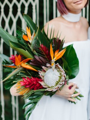 Papier peint  The bride in a dress with bare shoulders holds an exotic wedding bouquet of tropical flowers. Bouquet of protea, strelitzia, red ginger, and tropical leaves. Macramé in the background.