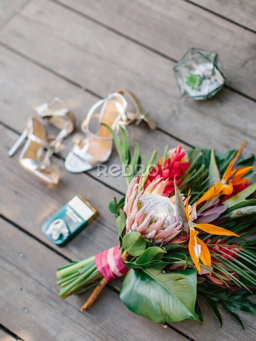Papier peint  The bride's bouquet in a tropical style with exotic flowers lies on a wooden floor. Bouquet of protea, palm leaves, strelitzia, and red ginger. In the background are women's shoes.
