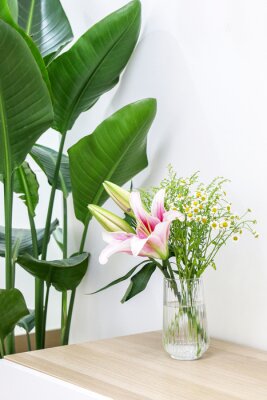 Papier peint  Vertical shot of bouquet of beautiful flowers (pink lilies and cute daises) in glass vase on wooden surface next to a Giant White Bird of Paradise (Strelitzia nicolai), decorating home interior