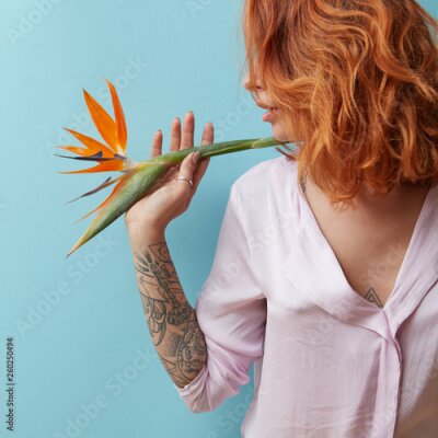 Papier peint  Woman with a tattoo holding an orange flower strelitzia in hands around a blue background with space for text. Valentine's day concept