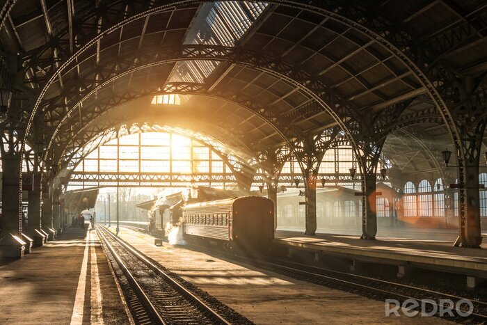 Poster  Ancienne gare avec un train et une locomotive sur le quai en attente de départ. Les rayons du soleil du soir dans les arcs de fumée.