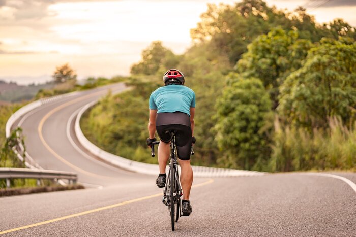 Poster  back view of a cyclist on top of a mountains winding road, riding a black bicycle down a hill, wearing bike helmet and blue cycling jersey, with grey clouds sunset sky and forest in the background.