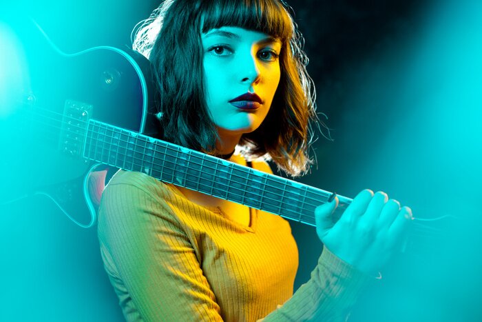 Poster  Beautiful young hipster woman with curly hair with red guitar in neon lights. Rock musician is playing electrical guitar. 90s style concept.