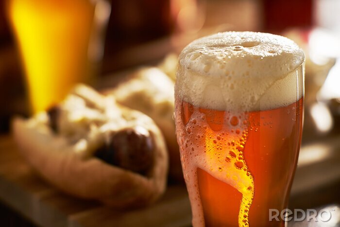 Poster  Bière IPA avec la tête écumeuse débordante dans la tasse servi avec bratwursts