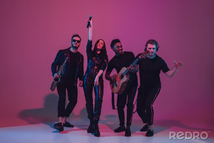 Poster  Group of young multiethnic musicians created band, dancing in neon light on pink background. Concept of music, hobby, festival, wellness. Joyful party host, dancer, singer, guitarist, saxophonist.