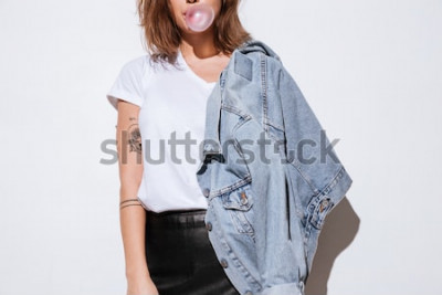 Poster  Photo recadrée de jeune femme vêtue d'une veste en jeans debout isolé sur fond blanc tout en soufflant des bulles avec du chewing-gum.