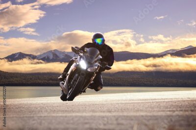 Poster  Un motard roule avec des montagnes en arrière-plan, parmi les nuages.