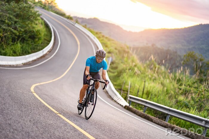 Sticker  asian male riding on a black bicycle along the winding road up a hill, wearing a cycling blue jersey, crash helmet and goggles, sunset light, grey sky, and forest trees and mountains in the background