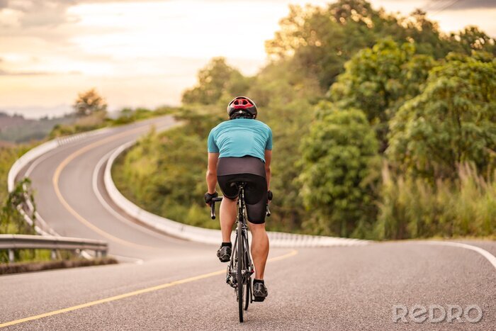 Sticker  back view of a cyclist on top of a mountains winding road, riding a black bicycle down a hill, wearing bike helmet and blue cycling jersey, with grey clouds sunset sky and forest in the background.