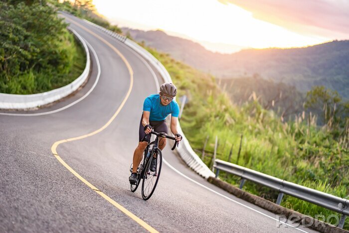 Tableau  asian male riding on a black bicycle along the winding road up a hill, wearing a cycling blue jersey, crash helmet and goggles, sunset light, grey sky, and forest trees and mountains in the background