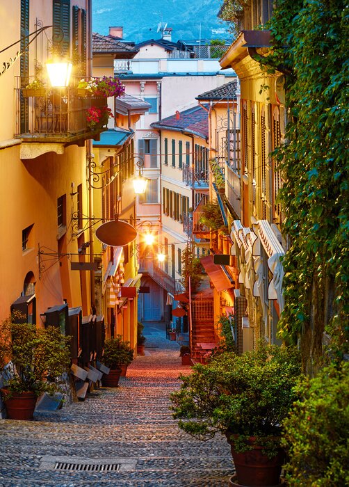 Tableau  Bellagio village at lake Como near Milan Italy, region Lombardy. Famous street with paving stones stairs and cosy restaurants during sunrise with glowing lanterns and green plants on old houses walls.
