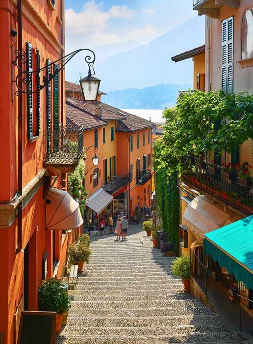 Tableau  Bellagio village at lake Como near Milan Italy, region Lombardy. Famous street with paving stones stairs and cosy restaurants during sunrise with glowing lanterns and green plants on old houses walls.