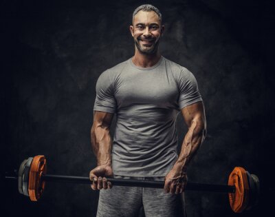 Tableau  Strong, adult, fit muscular caucasian man coach posing for a photoshoot in a dark studio under the spotlight wearing grey sportswear, showing his muscles and holding a sport gear looking confident and