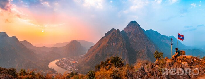 Tableau  Sunset panoramic view of couple of trekkers sitting on a rock on top of Nong Khiaw View Point with beautiful mountain and Nam Ou river in background, Luang Prabang Province, Northern Laos.