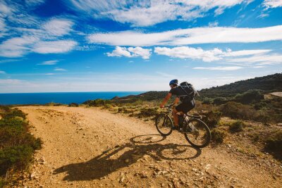 Tableau  un jeune mec monté sur un vélo de montagne sur une route cyclable en Espagne sur la route dans le contexte de la mer Méditerranée. Habillé dans un casque, un noir et un sac à dos noir