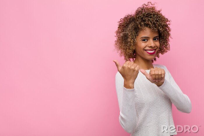 Tableau  young african american woman smiling cheerfully and casually pointing to copy space on the side, feeling happy and satisfied against pink wall